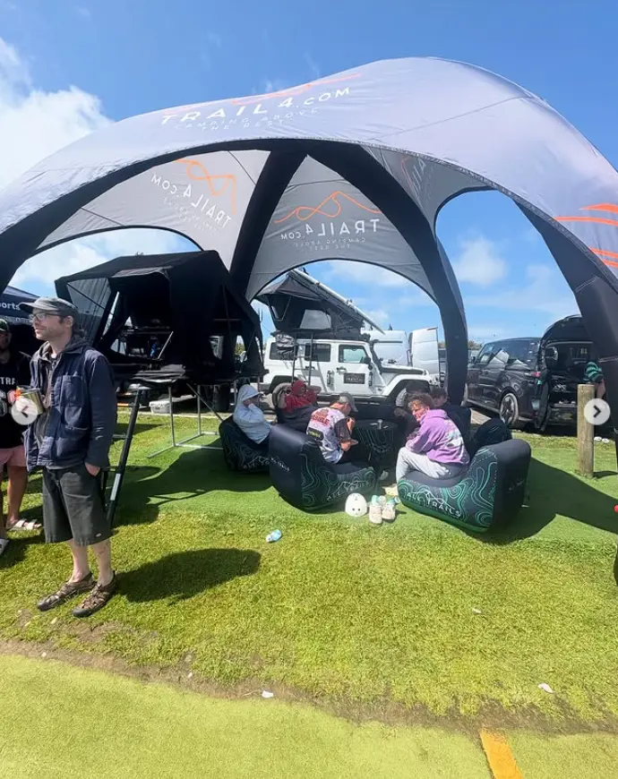 A group of people hanging out and socializing under a large grey and black branded inflatable dome tent, with a rooftop tent on display and attendees sitting on inflatable chairs during a daytime outdoor gathering.